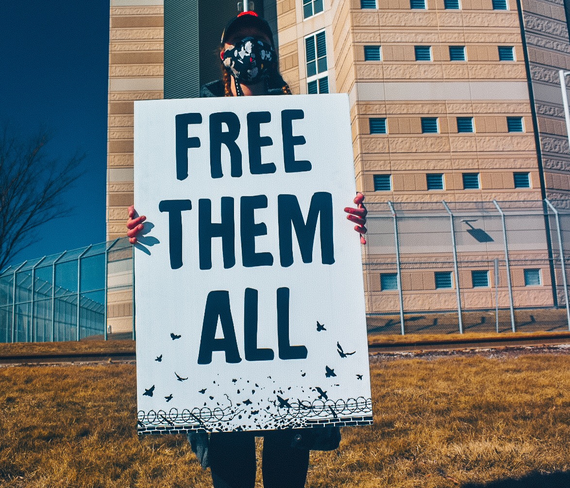 picture of a protester holding a placard saying 'free them all' with the Toronto Prisoner's Rights Project logo underneath